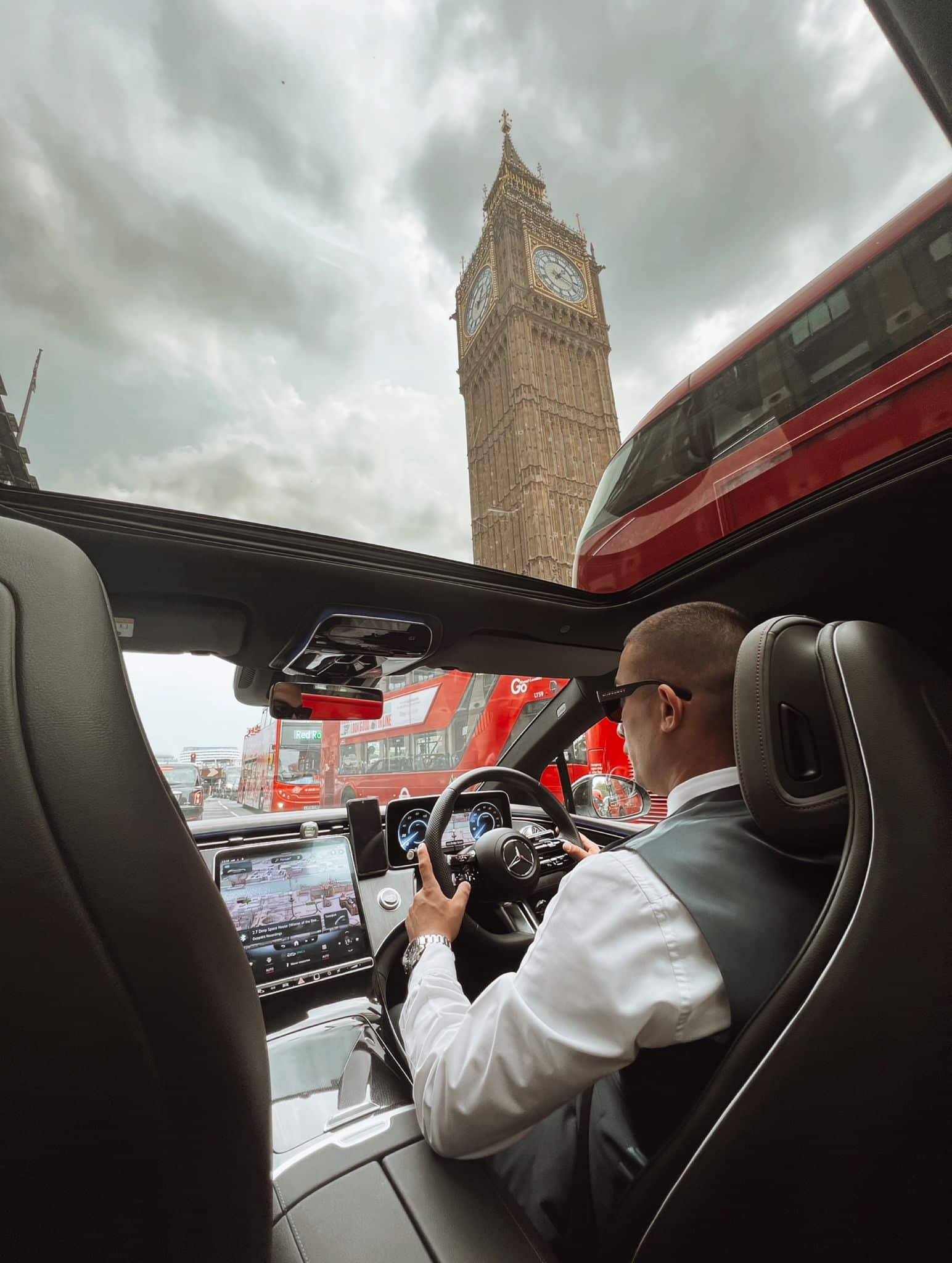 A man driving a car with Big Ben clock tower prominently visible in the background