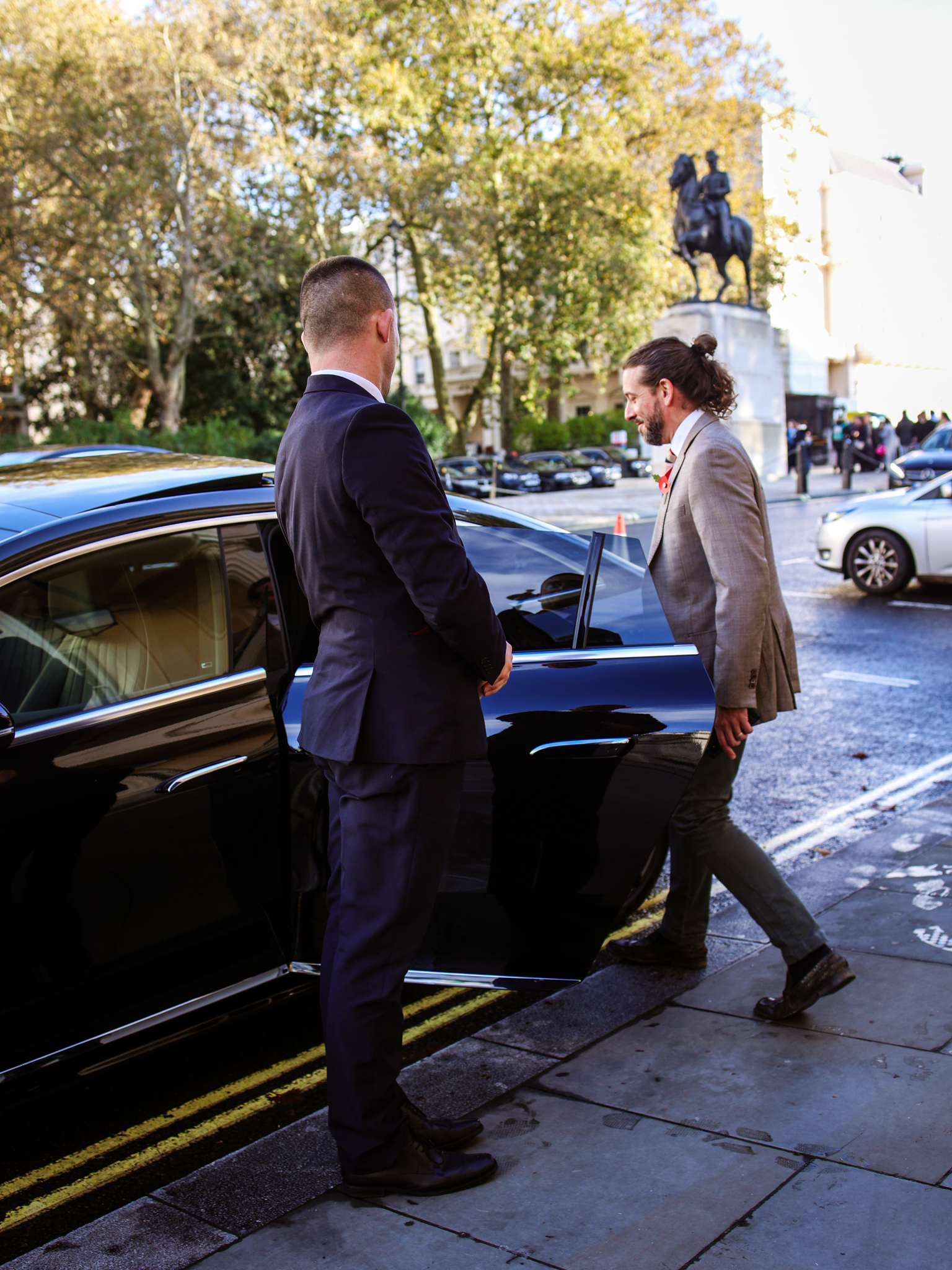 A man stepping in of a black car, wearing casual attire, with a city street in the background.